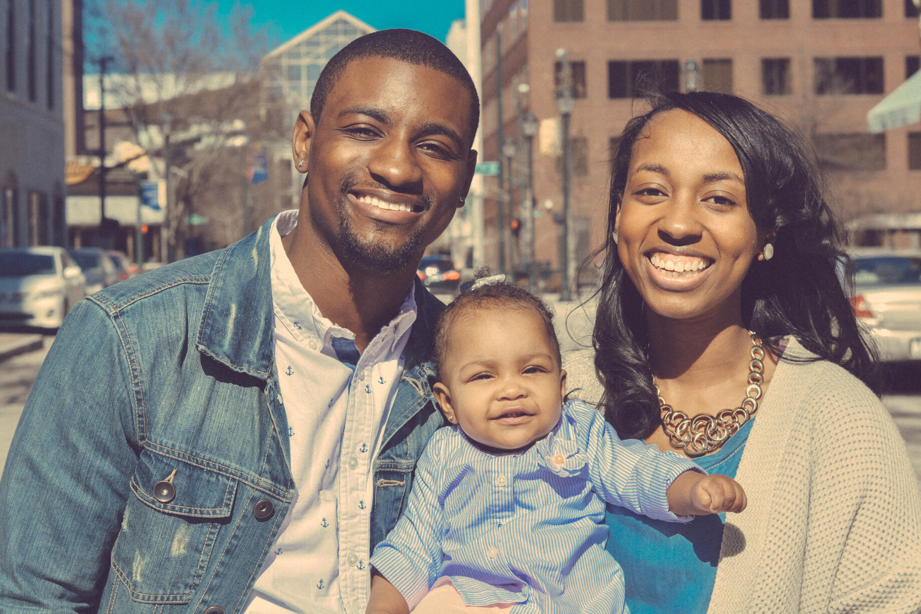 Two smiling adults and a young infant pose on a sidewalk