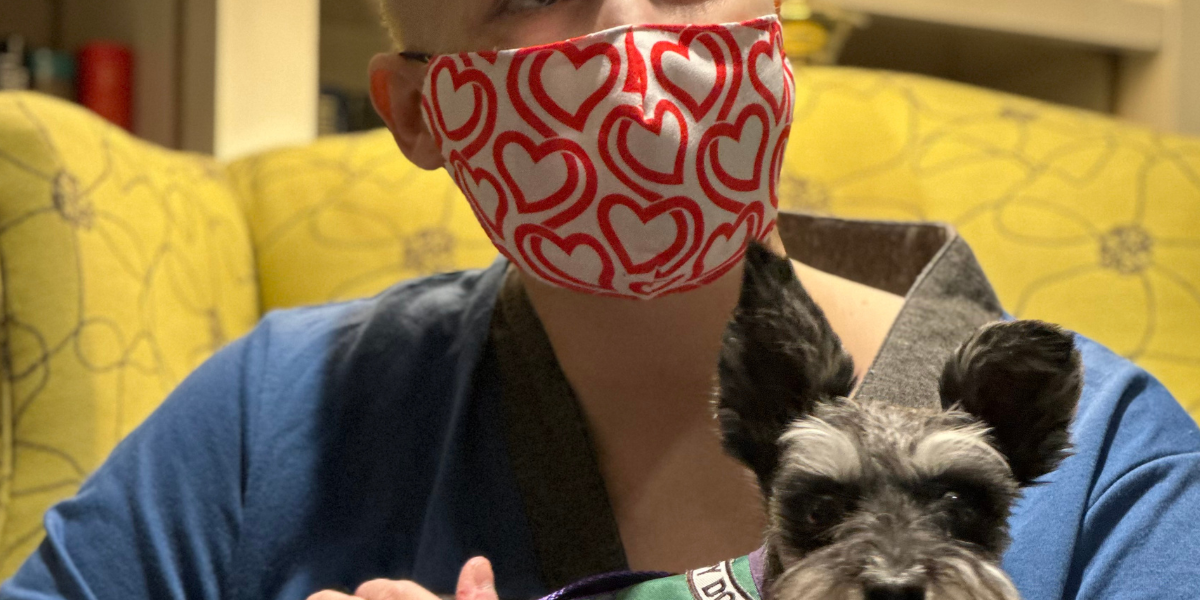 An adult wearing a mask sits in a library with a miniature schnauzer dog on her lap