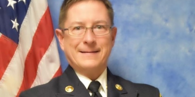 A smiling adult wears a firefighter suitcoat in front of a U-S-A flag