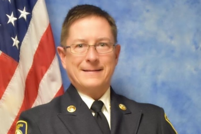 A smiling adult wears a firefighter suitcoat in front of a U-S-A flag